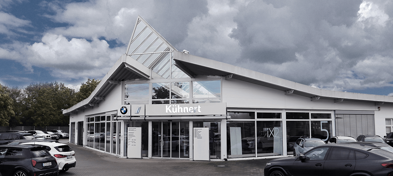 Außenansicht des Autohauses Kühnert in Altenburg mit moderner Glasfront und schrägem Dach. Vor dem Gebäude sind mehrere Autos geparkt, das BMW-Logo und der Kühnert-Schriftzug sind sichtbar. Im Hintergrund ein bewölkter Himmel.