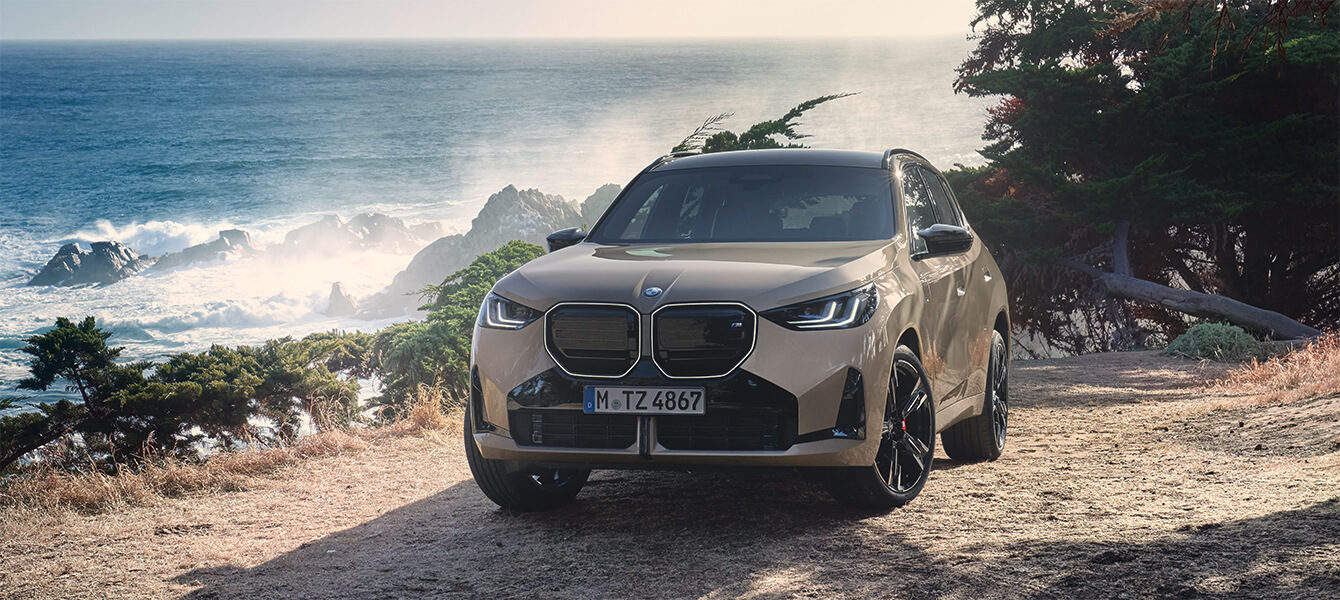 BMW X3 SUV auf einer felsigen Küstenstraße mit Blick auf das Meer und Wellen im Hintergrund.