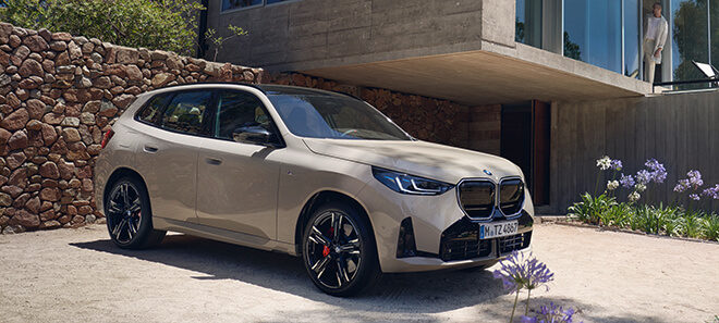 Ein BMW X3 in Beige, geparkt vor einem modernen Haus mit einer Steinmauer und Gartenpflanzen im Vordergrund.