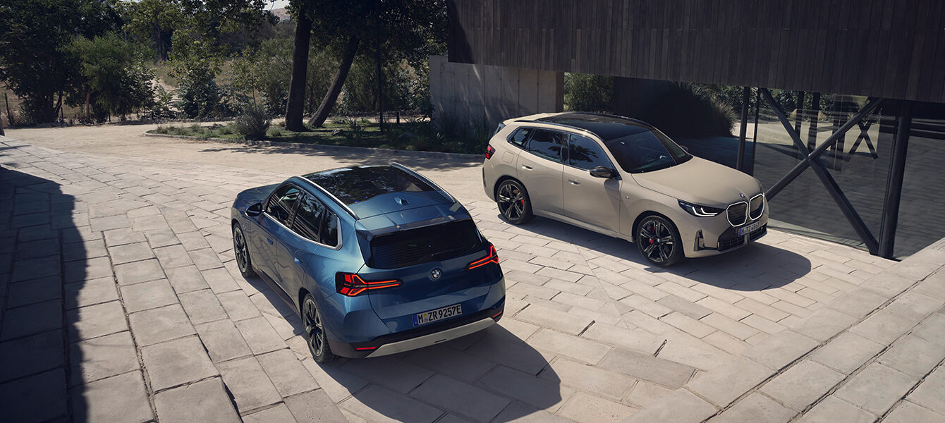 Zwei BMW X3 Modelle, eins in Blau und eins in Beige, parken vor einem modernen Gebäude auf einer gepflasterten Fläche.