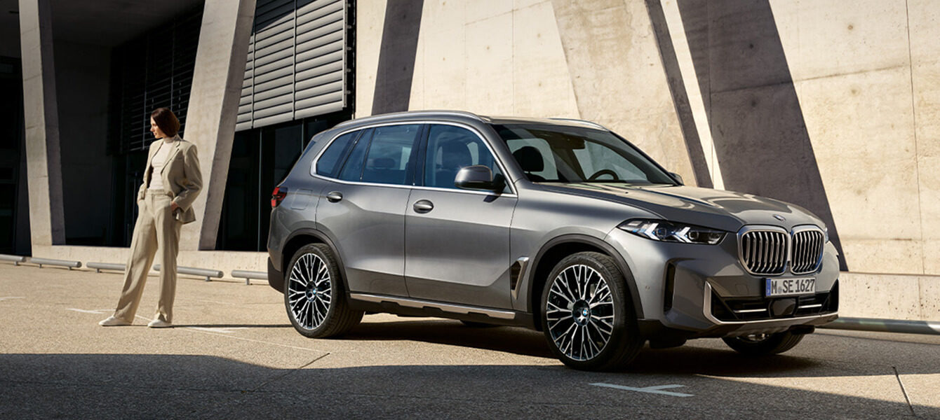 Grauer BMW X5 geparkt vor einer modernen Betonfassade, mit einer Frau in beiger Kleidung, die in der Nähe des Fahrzeugs steht.