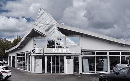 Außenansicht des Autohauses Kühnert in Altenburg mit moderner Glasfront und schrägem Dach. Vor dem Gebäude sind mehrere Autos geparkt, das BMW-Logo und der Kühnert-Schriftzug sind sichtbar. Im Hintergrund ein bewölkter Himmel.